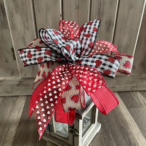 May include: A white lantern with a red, black, and white ribbon bow on top. The ribbon has a heart pattern and is tied in a bow. The lantern is sitting on a wooden surface.
