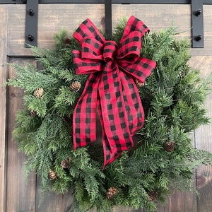 May include: A Christmas wreath featuring a lush arrangement of green pine needles and small pine cones. A large red and black buffalo plaid bow is centered at the top. The wreath is displayed against a dark wooden door.