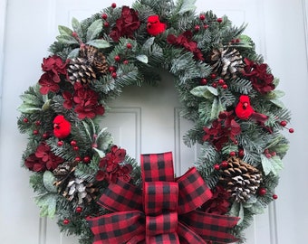 May include: A Christmas wreath with red cardinals, red berries, pine cones, and a red and black buffalo plaid bow.