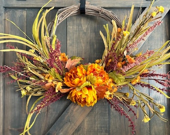 May include: A fall-themed wreath with a grapevine base, adorned with vibrant orange and yellow flowers, burgundy accents, and green foliage. The wreath is displayed on a dark wooden door, creating a warm and inviting seasonal decoration.