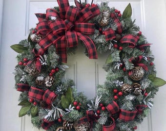 May include: A 24-inch wreath featuring artificial evergreen branches with a light dusting of white flocking. The wreath is decorated with red and black buffalo plaid ribbon, clusters of red berries, natural pinecones, and green leaves.