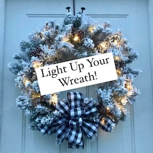 May include: A snow-covered wreath with pinecones and warm white lights, adorned with a black and white buffalo check bow. The wreath hangs on a white door and features the text "Light Up Your Wreath!"