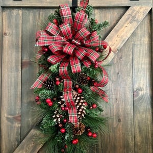May include: A Christmas swag featuring faux pine needles, red berries, and pine cones. A large red and green plaid bow is the focal point. The swag is designed to hang on a door or wall, adding festive holiday decor.