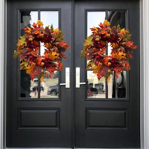 May include: Two identical circular wreaths featuring a dense arrangement of artificial autumn foliage in shades of orange, red, yellow, and brown, accented with small berries and pinecones, mounted on a black double door with glass panels.