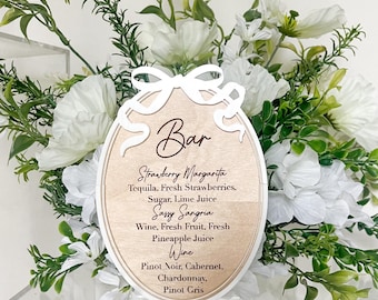 May include: A wooden sign with a white bow and the word "Bar" written in cursive. The sign lists drink options including Strawberry Margarita, Sassy Sangria, and Wine.