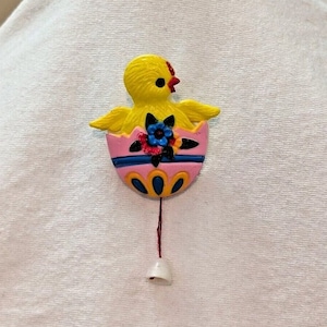 May include: A yellow chick with a red beak peeks out of a pink and blue Easter egg with yellow and blue accents. The egg has a string attached to a white bell.