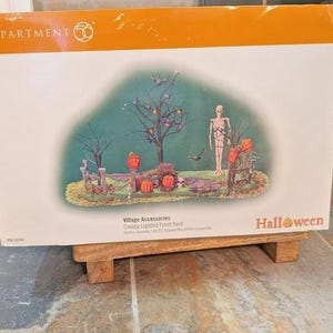 May include: A Halloween-themed display box featuring a spooky front yard scene. The illustration includes a skeleton, pumpkins, a tree with bats, and a fence. The box is labeled "Village Accessories" and "Creepy Lighted Front Yard."