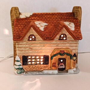 May include: A ceramic cottage with a brown roof and white snow accents. The cottage has a lit window and a sign that reads "Cracker Cottage".