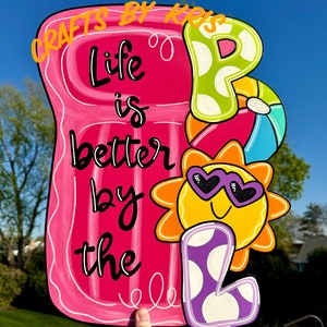 May include: A pink pool float with the text "Life is better by the pool" in black. A purple letter "L" with white polka dots is next to a yellow sun with sunglasses. The sun is behind a green letter "P" with white polka dots and a blue and white beach ball.
