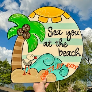 May include: A wooden sign with a beach theme. It features a palm tree, a sun, a wave, a seagull, and the text "Sea you at the beach". The sign is painted in bright colors and has a whimsical style.