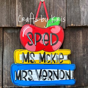 May include: A red apple with white polka dots and the word "SPED" on it, stacked on top of two books. The top book is yellow with the text "MS. McKIE" and the bottom book is blue with the text "MRS. VERNON".