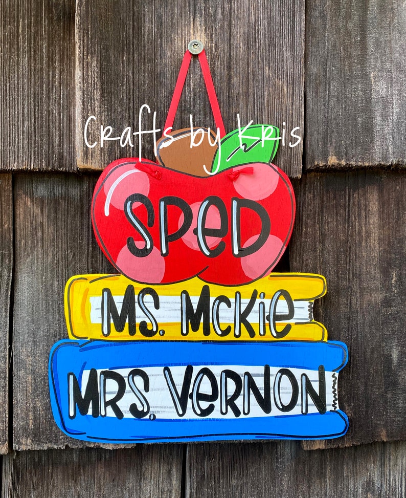May include: A red apple with white polka dots and the word "SPED" on it, stacked on top of two books. The top book is yellow with the text "MS. McKIE" and the bottom book is blue with the text "MRS. VERNON".
