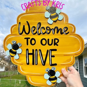 May include: A wooden, yellow, beehive-shaped sign with the words "Welcome to Our Hive" in black script. Three cartoon bees with black and yellow bodies and light blue wings are attached to the sign. The sign is held by a hand.
