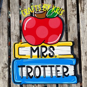 May include: A red apple with white polka dots and a brown stem sits on top of two stacked books. The top book is yellow with black text that reads "MRS.". The bottom book is blue with black text that reads "TROTTER". The text "CRAFTS BY KRIS" is written in yellow above the apple.