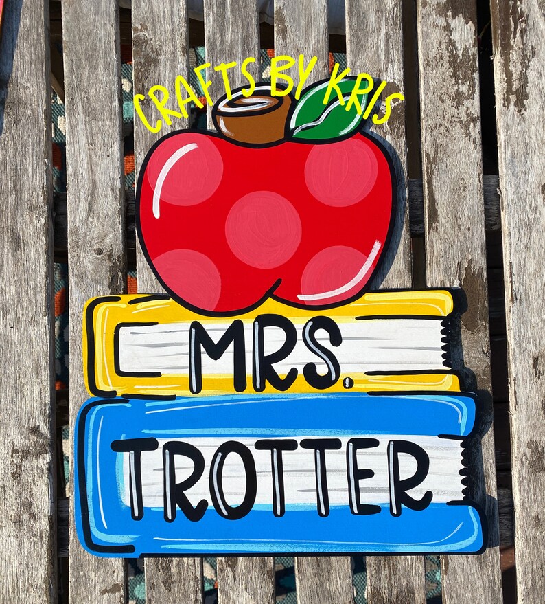 May include: A red apple with white polka dots and a brown stem sits on top of two stacked books. The top book is yellow with black text that reads "MRS.". The bottom book is blue with black text that reads "TROTTER". The text "CRAFTS BY KRIS" is written in yellow above the apple.
