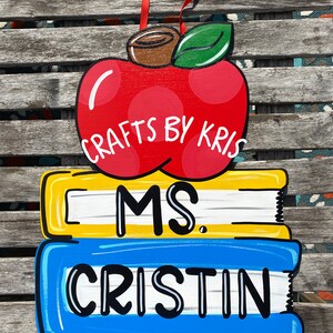 May include: A red apple with a green leaf and the text "CRAFTS BY KRIS" on top of two stacked books. The top book is yellow with white stripes and the text "MS." The bottom book is blue with white stripes and the text "CRISTIN".
