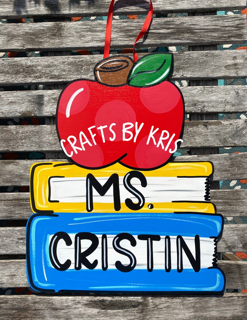May include: A red apple with a green leaf and the text "CRAFTS BY KRIS" on top of two stacked books. The top book is yellow with white stripes and the text "MS." The bottom book is blue with white stripes and the text "CRISTIN".