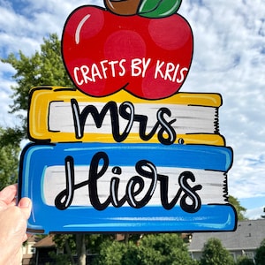 May include: A wooden sign with a red apple, three stacked books, and the text "Mrs. Liers" in black cursive. The apple has white polka dots and a green leaf. The books are yellow, blue, and white. The sign says "Crafts by Kris" at the top.