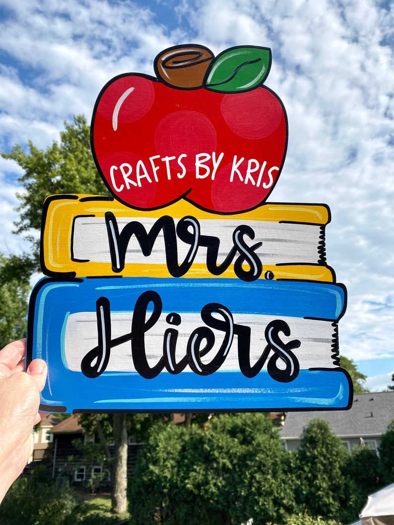 May include: A wooden sign with a red apple, three stacked books, and the text "Mrs. Liers" in black cursive. The apple has white polka dots and a green leaf. The books are yellow, blue, and white. The sign says "Crafts by Kris" at the top.