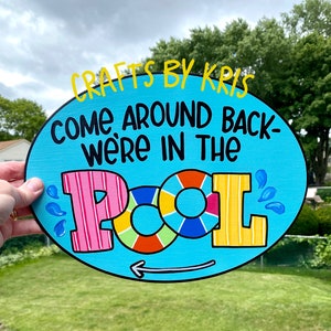 May include: A blue and white oval sign with the text "Crafts by Kris Come Around Back - We're in the Pool" in black lettering. The word "Pool" is in large, colorful letters with a life preserver in the middle.