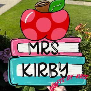 May include: A wooden sign with a pink apple on top of two stacked books. The top book is pink and says "MRS." The bottom book is teal and says "KIRBY".