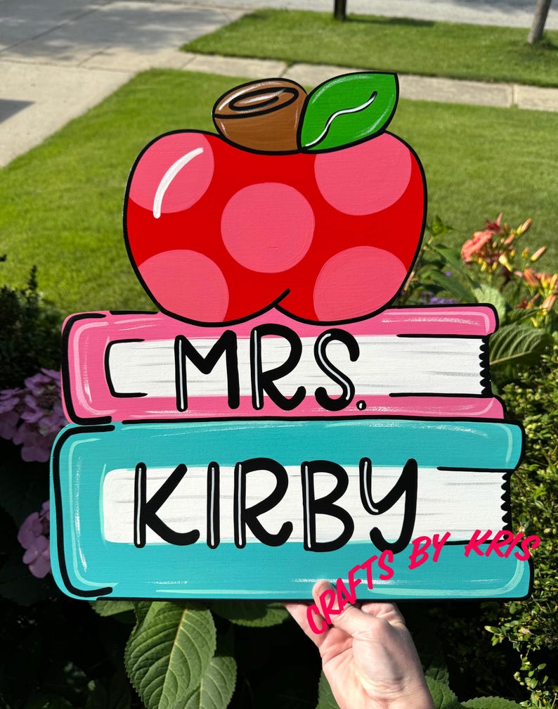 May include: A wooden sign with a pink apple on top of two stacked books. The top book is pink and says "MRS." The bottom book is teal and says "KIRBY".