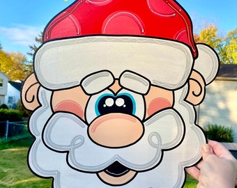 May include: A white and red Santa Claus face cutout with a white beard and blue eyes. The cutout is held in a hand against a blue sky background. The text "CRAFTS BY KRIS" is written in yellow letters at the top of the image.