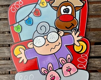 May include: A red truck with a white trim and a gray bed. A woman with gray hair and glasses is sitting in the bed of the truck wearing a purple sweater and pink bunny slippers. A reindeer with brown fur and a red nose is sitting in the passenger seat. The truck is decorated with a string of colorful lights.