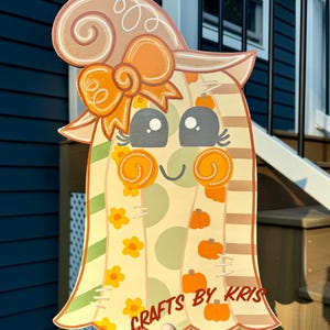 May include: A wooden cutout of a ghost wearing a witch hat, decorated with orange bows, yellow flowers, and small pumpkins. The ghost features large gray eyes, rosy cheeks, and the text CRAFTS BY KRIS printed in brown at the bottom.