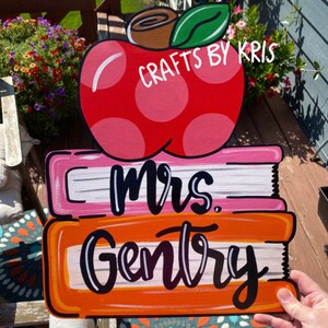 May include: A red apple with white polka dots and a green leaf sits on top of two stacked books. The top book is pink with white stripes and the bottom book is orange with white stripes. The words "Mrs. Gentry" are written in black on the books.