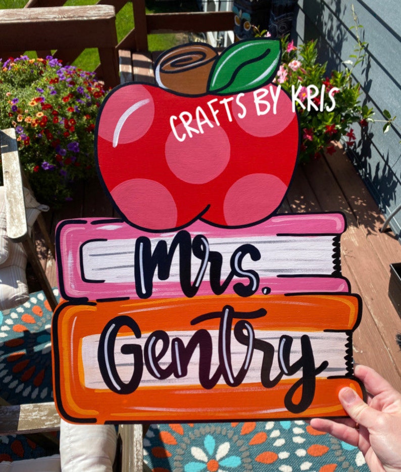 May include: A red apple with white polka dots and a green leaf sits on top of two stacked books. The top book is pink with white stripes and the bottom book is orange with white stripes. The words "Mrs. Gentry" are written in black on the books.