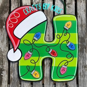 May include: A wooden Christmas decoration in the shape of the letter "H". The letter is painted green with horizontal stripes and decorated with colorful Christmas lights. A red Santa hat is on the top left. The text "Crafts by Kris" is written on the top.