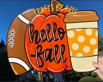 May include: Wooden fall-themed sign featuring a football, pumpkin, and coffee cup. The pumpkin is orange with the words "hello fall" in black script. The coffee cup has an orange polka dot design.