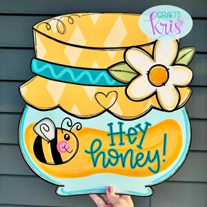 May include: A yellow and blue honey jar door hanger with a bee and a daisy. The jar has a yellow lid with a blue ribbon and the text "Hey honey!" is written on the front.
