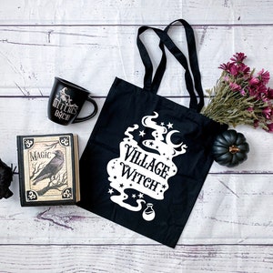 May include: Black tote bag with white text that reads "Village Witch" and a graphic of a cauldron with smoke and stars. The bag is on a white wooden surface with a black mug, a book with a raven on the cover, a black raven figurine, and a small purple flower bouquet.