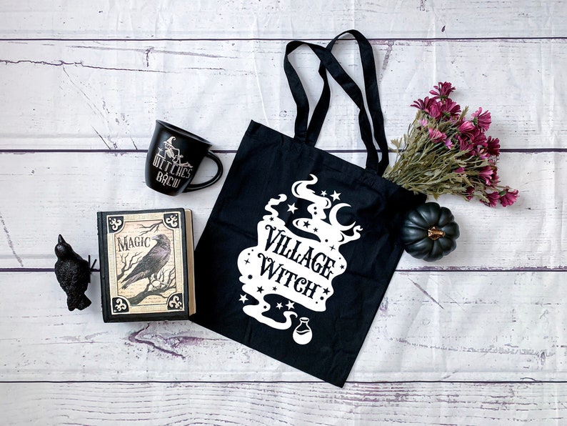 May include: Black tote bag with white text that reads "Village Witch" and a graphic of a cauldron with smoke and stars. The bag is on a white wooden surface with a black mug, a book with a raven on the cover, a black raven figurine, and a small purple flower bouquet.