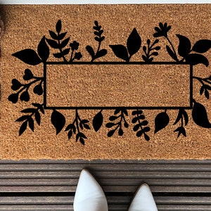 May include: A brown coir doormat with a black floral design and a rectangular space for text. The doormat is surrounded by fall foliage.