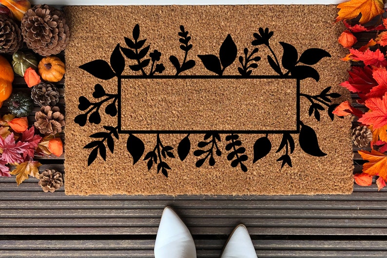 May include: A brown coir doormat with a black floral design and a rectangular space for text. The doormat is surrounded by fall foliage.