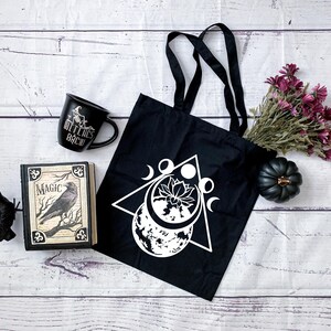 May include: Black tote bag with a white design of a triangle, moon phases, and a lotus flower. The bag is on a white wooden surface with a black mug that says "Witches Brew", a book titled "Magic", and a black crow figurine.