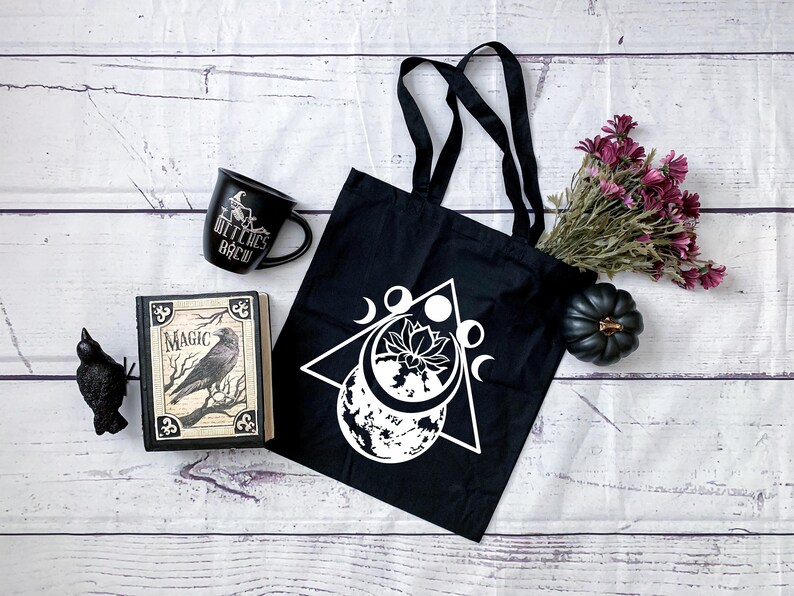 May include: Black tote bag with a white design of a triangle, moon phases, and a lotus flower. The bag is on a white wooden surface with a black mug that says "Witches Brew", a book titled "Magic", and a black crow figurine.