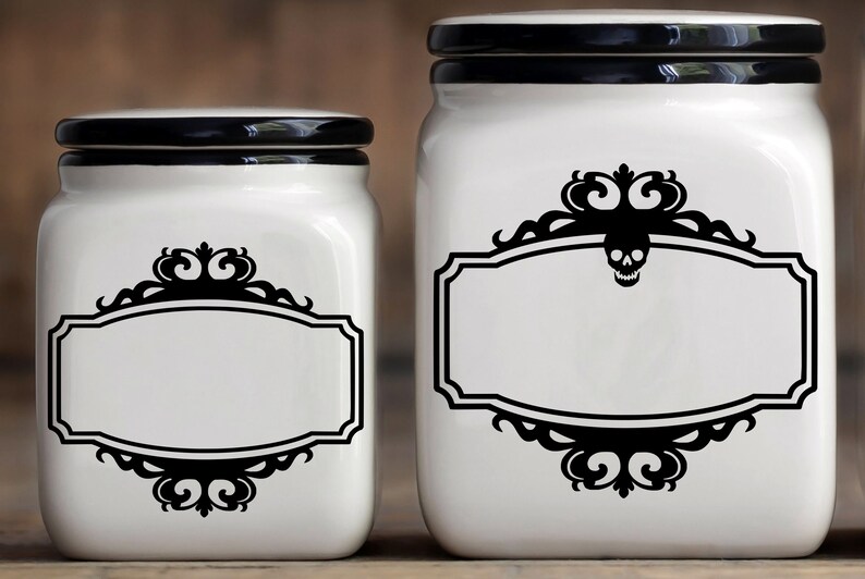 May include: Two white ceramic canisters with black lids and black decorative labels featuring a skull and ornate scrollwork. The labels have blank spaces for writing.