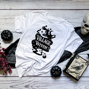 May include: White t-shirt featuring a black graphic that reads "Village Witch" with a crescent moon, stars, and a potion bottle. The shirt is displayed on a white wooden surface with black fabric, a black book with a raven, and black pumpkins.