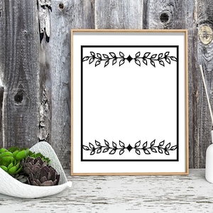 May include: A black and white framed print with a simple leaf design border. The print is on a white background and the frame is a light wood color. The print is sitting on a wooden surface.