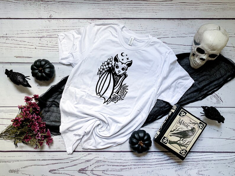 Op de afbeelding: Wit T-shirt met een zwart-witte afbeelding van een vleermuis met bloemen en een halve maan. Het shirt ligt op een witte houten achtergrond met zwarte stof, een schedel en een boek met het woord "Magic" op de omslag.