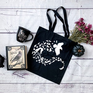 May include: Black tote bag with a white design of a raven, stars, moons, and other magical symbols. The bag is on a white wooden surface with a book, a mug that says "Witches Brew", a black raven figurine, and a bouquet of purple flowers.