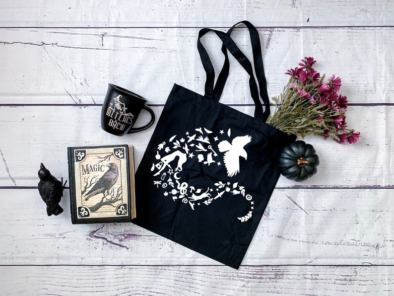 May include: Black tote bag with a white design of a raven, stars, moons, and other magical symbols. The bag is on a white wooden surface with a book, a mug that says "Witches Brew", a black raven figurine, and a bouquet of purple flowers.