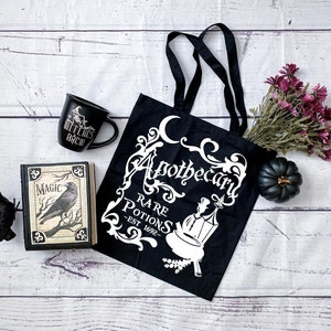 May include: Black tote bag with white text that reads "Apothecary Rare Potions - Est 1692" with a crescent moon and floral design. A black mug with white text that reads "Witches Brew" and a black book with white text that reads "Magic" are also in the image.