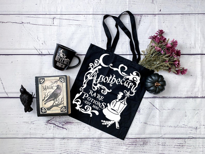 May include: Black tote bag with white text that reads "Apothecary Rare Potions - Est 1692" with a crescent moon and floral design. A black mug with white text that reads "Witches Brew" and a black book with white text that reads "Magic" are also in the image.