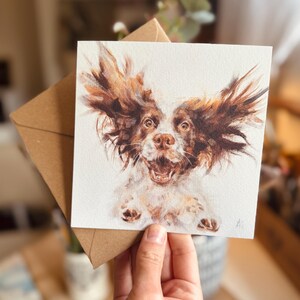 Op de afbeelding: Vierkante wenskaart met een aquarel schilderij van een hond met bruin en wit haar, de oren omhoog. De bek van de hond staat open in een vrolijke uitdrukking. De kaart wordt voor een bruine envelop gehouden.