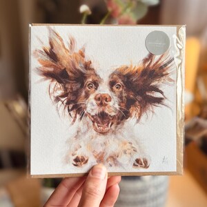 Op de afbeelding: Een vierkante wenskaart met een aquarel schilderij van een hond met bruin en wit haar, zijn oren wapperen. De hond heeft een open mond en lijkt te glimlachen. De kaart zit op een bruine achtergrond en heeft een kleine, ronde sticker.